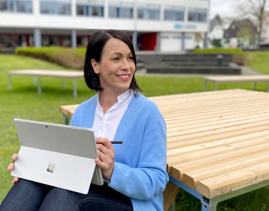 Kerstin Eller sitzt mit einem Tablet in der Hand auf einer Bank vor einer Schule.